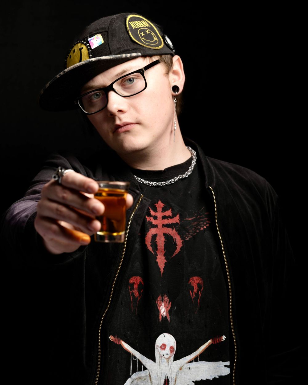 Young man in black cap and glasses holding a shot glass, wearing a dark graphic tee with red skull design.