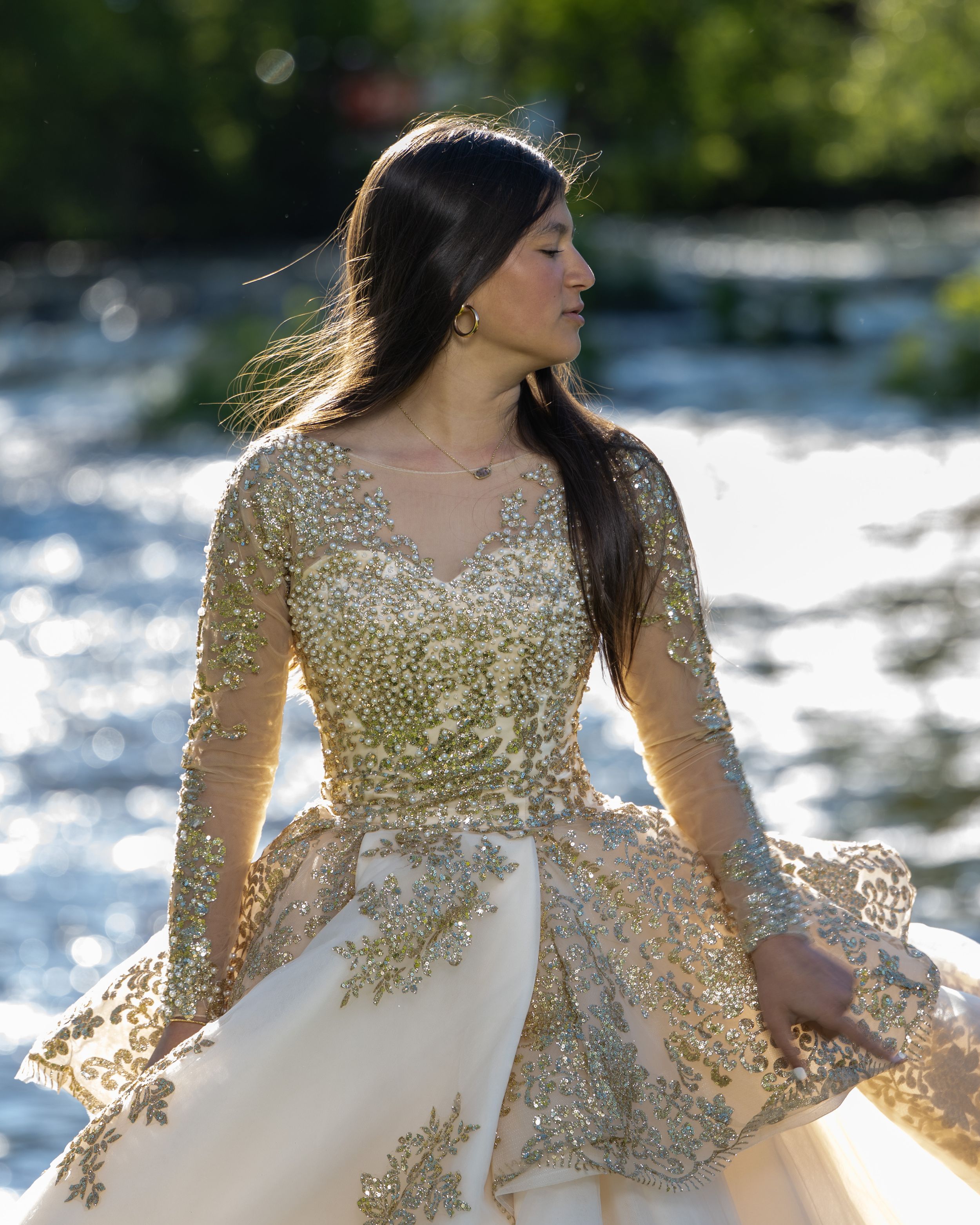 Woman in gold sequined ball gown poses gracefully by a sparkling river in natural sunlight.