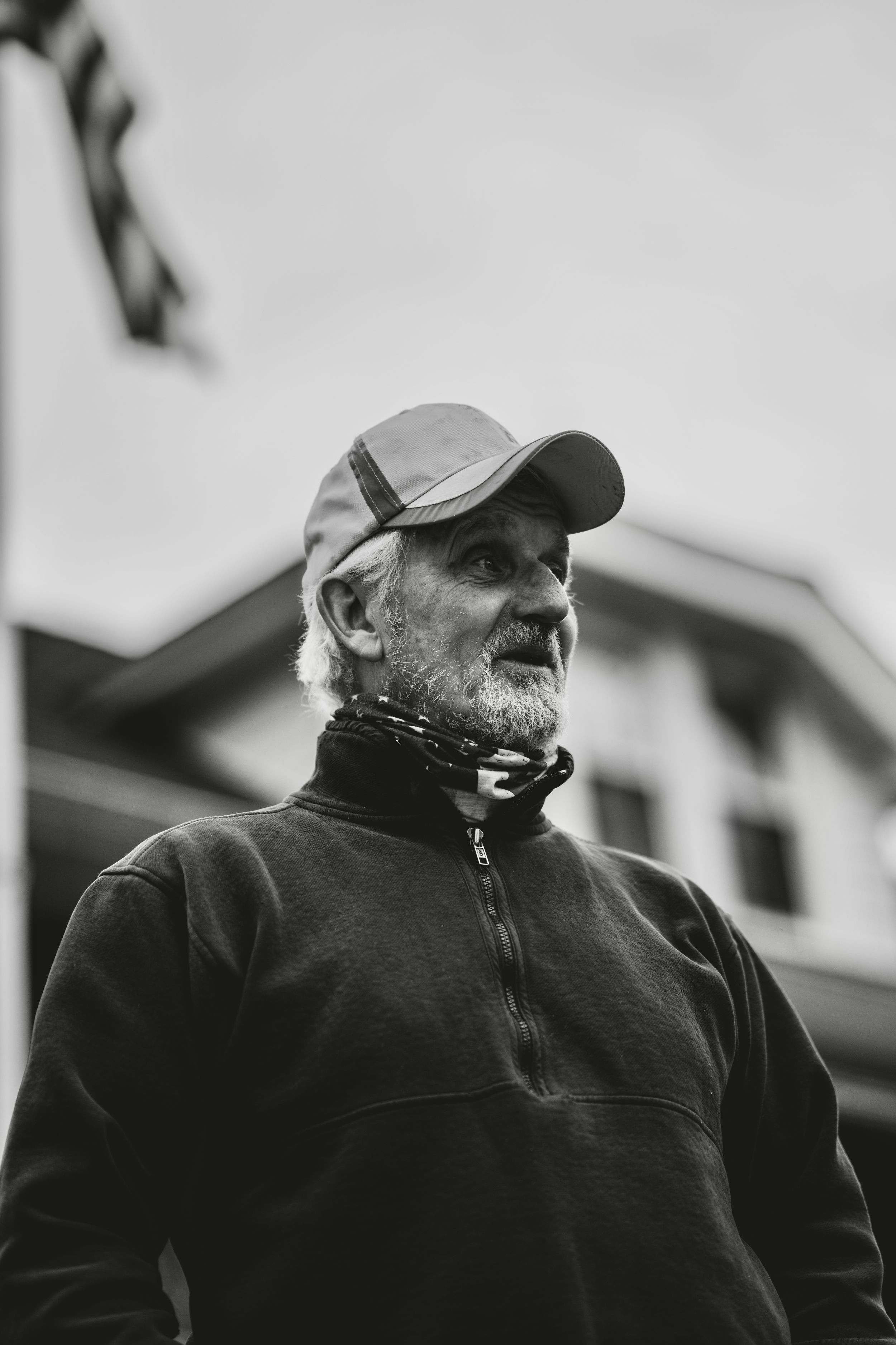 Black and white portrait of an elderly bearded man wearing a cap and neck gaiter outdoors.