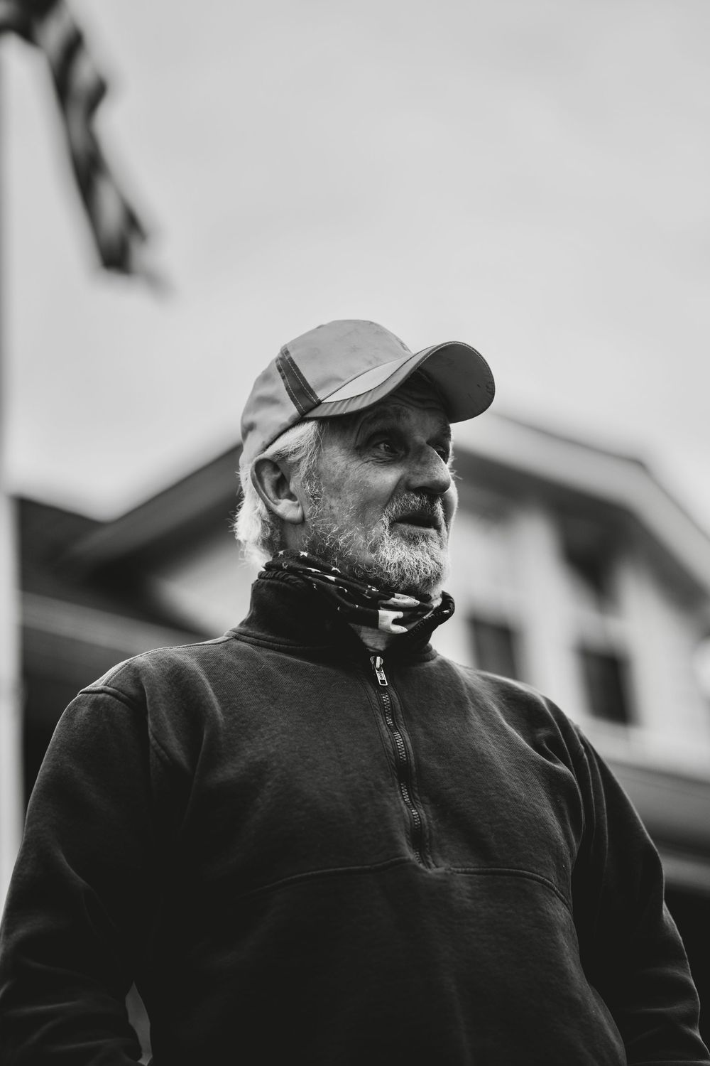 Black and white portrait of an elderly bearded man wearing a cap and neck gaiter outdoors.