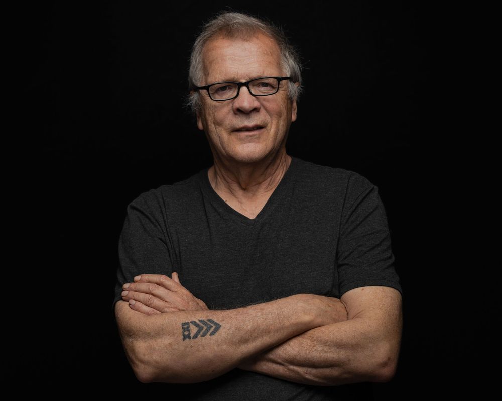 Older man with glasses and arm tattoo, arms crossed, wearing black V-neck shirt, posing against dark background.