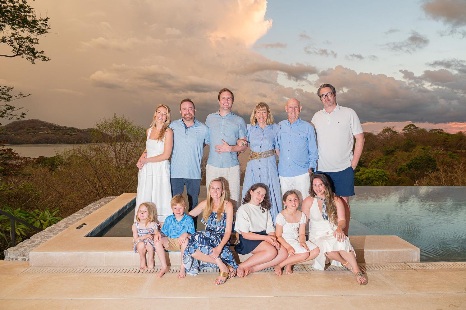 Large family group portrait at sunset by infinity pool with tropical landscape and dramatic sky.