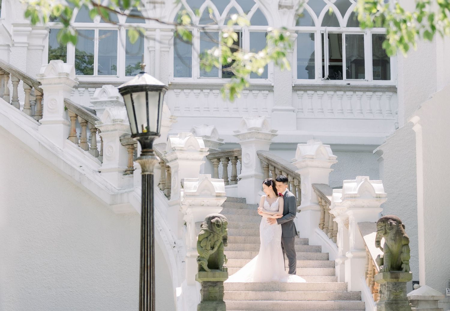 A bride and groom embrace on elegant white marble stairs outside a grand white wedding venue framed by green trees.