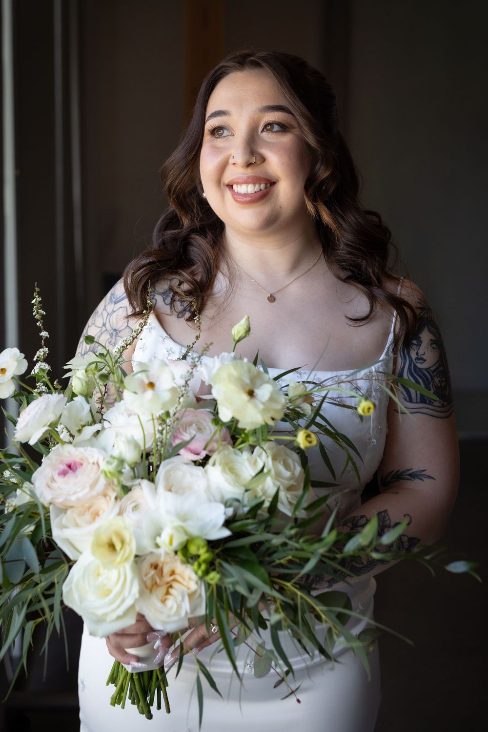 Radiant bride with tattoos smiles holding large white and blush floral bouquet in soft indoor light.