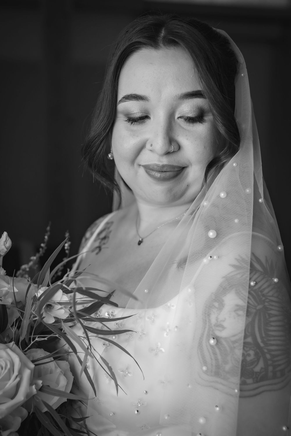 Close-up black and white portrait of bride in pearl veil smiling softly while holding bouquet.