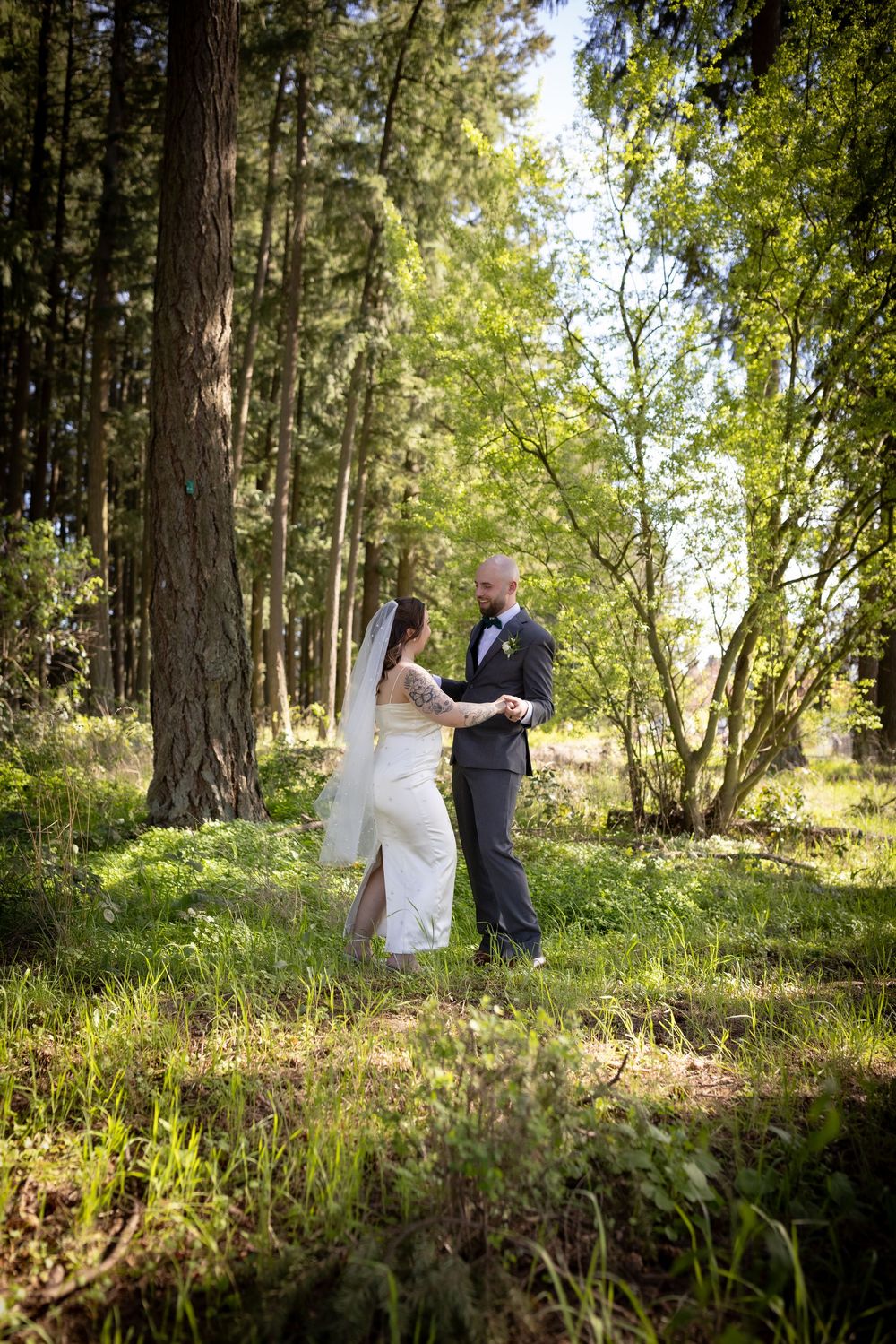 Bride and groom share a romantic embrace surrounded by lush green trees in sunlit forest setting.