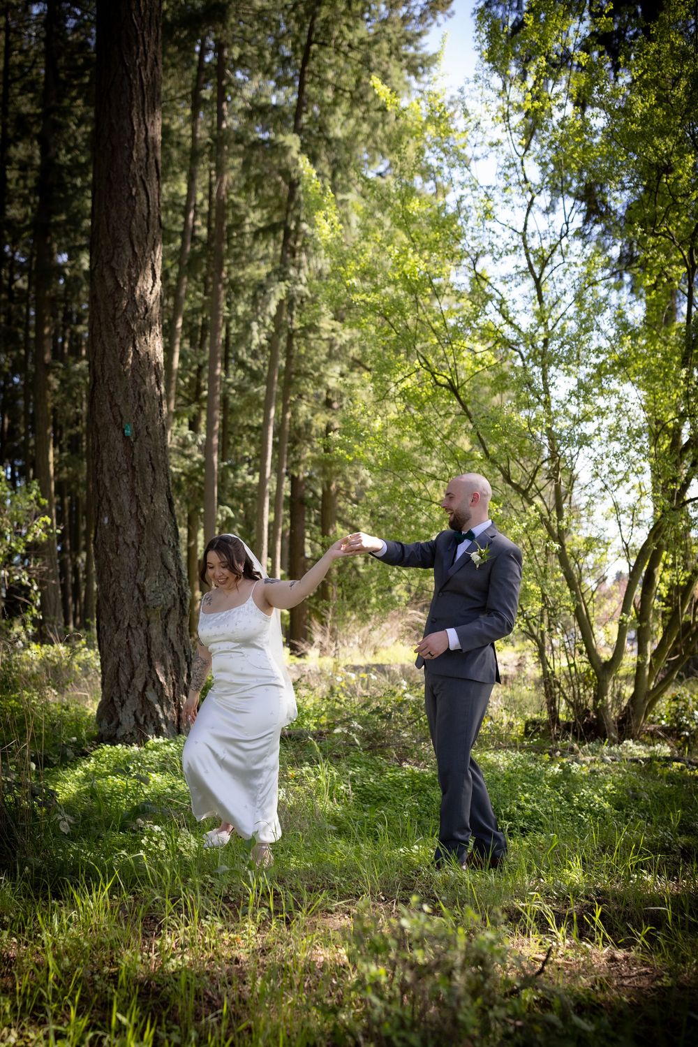 Bride and groom dance joyfully among tall trees in sunlit forest clearing on wedding day.