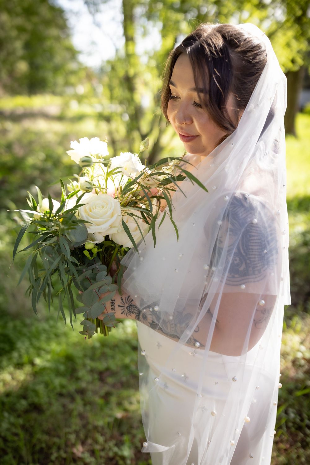 Bride in white gown and veil holds bouquet of white roses and greenery outdoors.