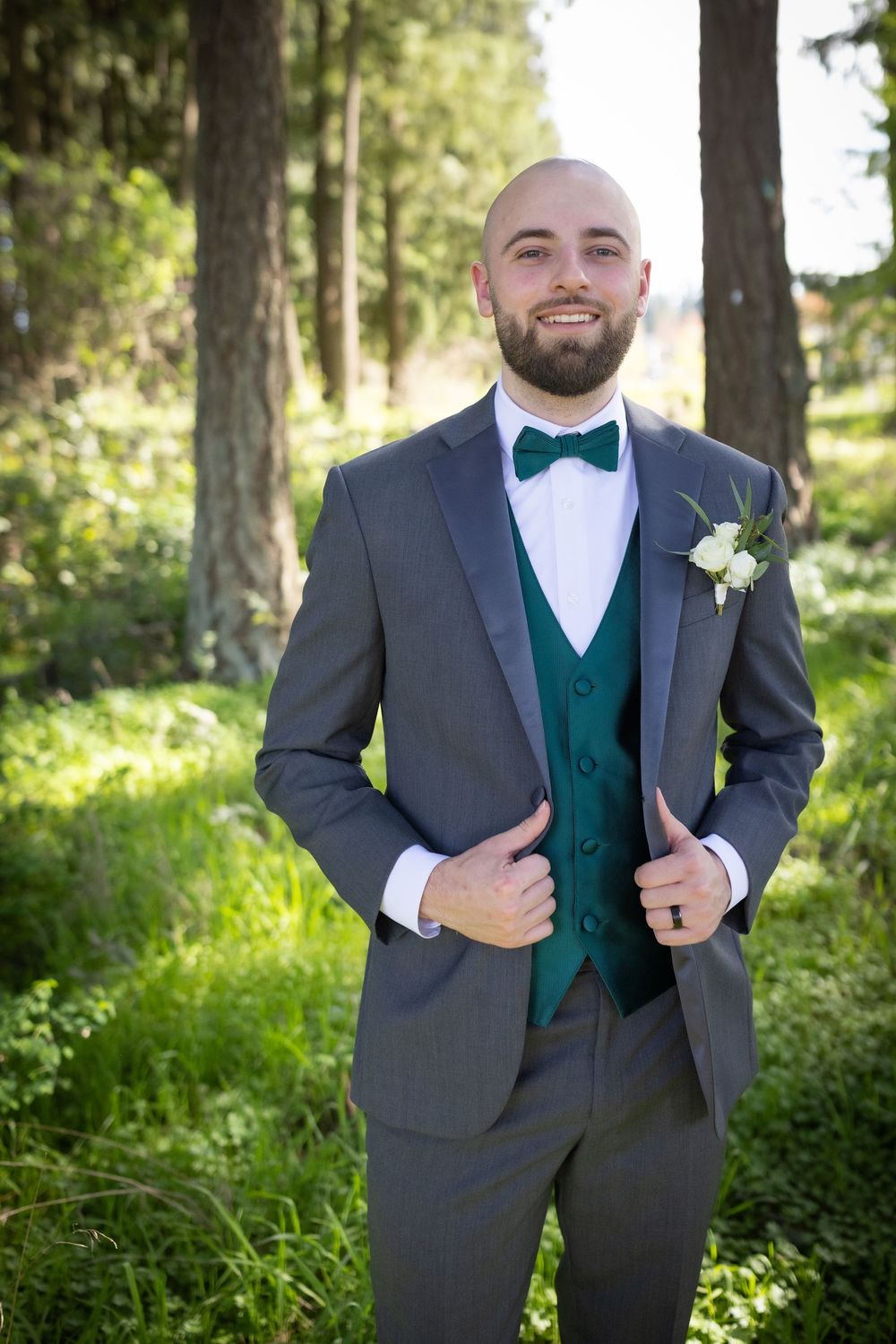 Groom in gray suit with green vest and bow tie smiles in a forest setting.