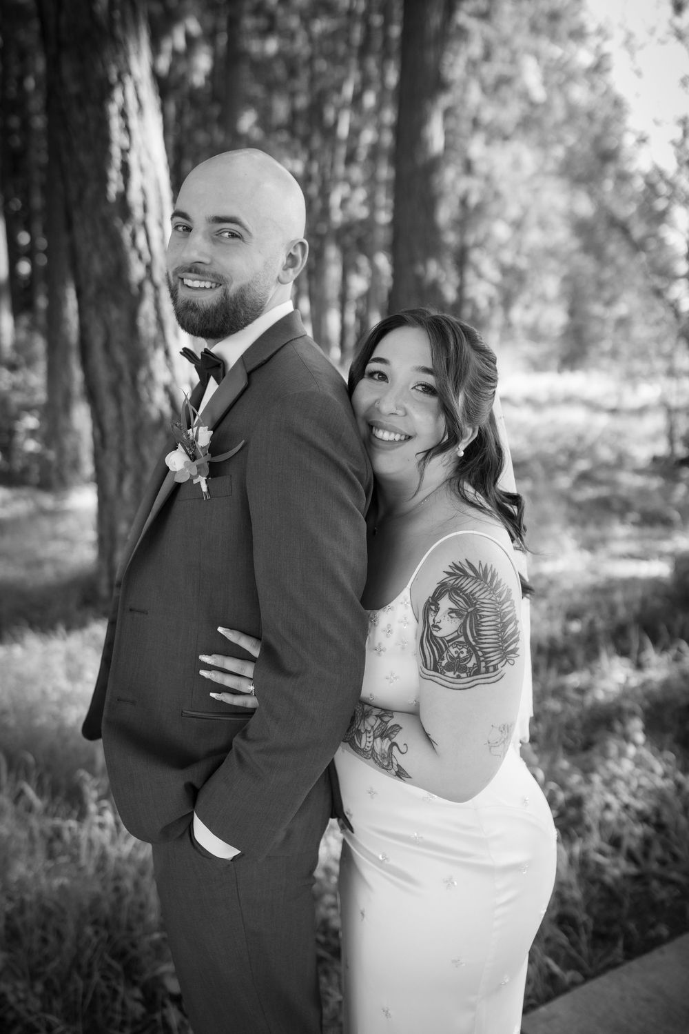 Black and white photo of tattooed bride and groom posing back-to-back in forest.