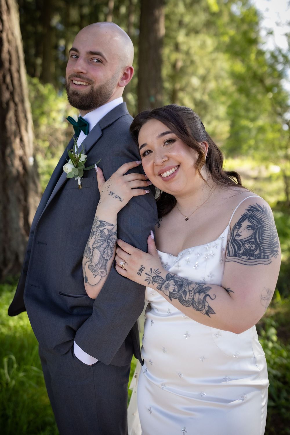 Tattooed bride in white gown leans on groom in gray suit outdoors in forest.