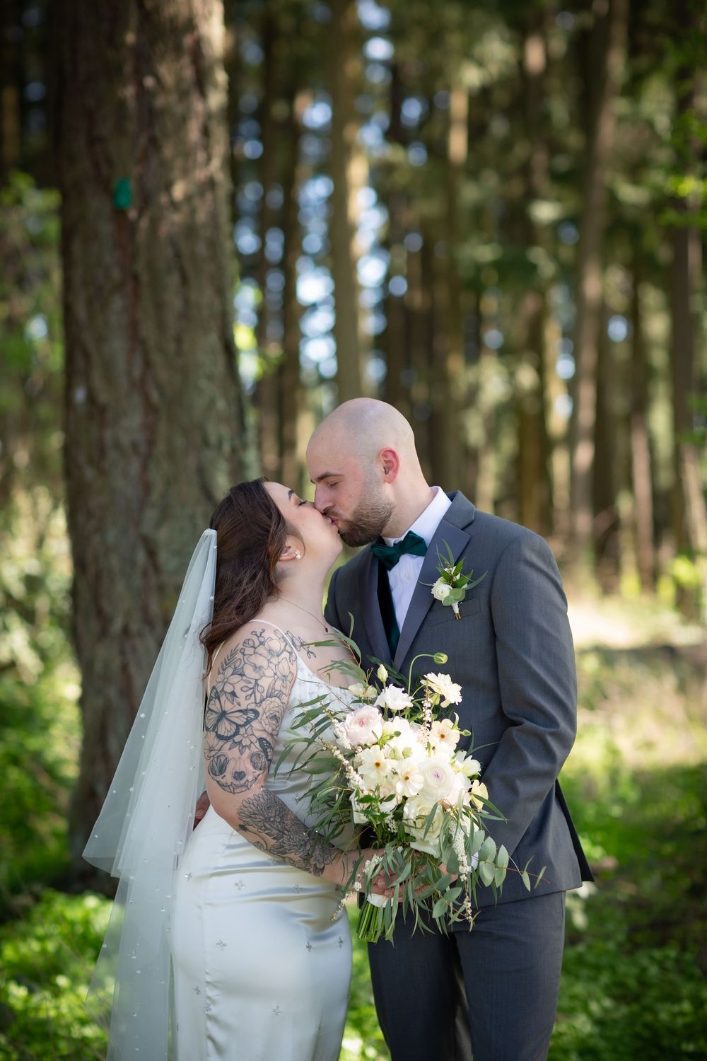 Bride and groom share a kiss in a sunlit forest, bride holding white bouquet.