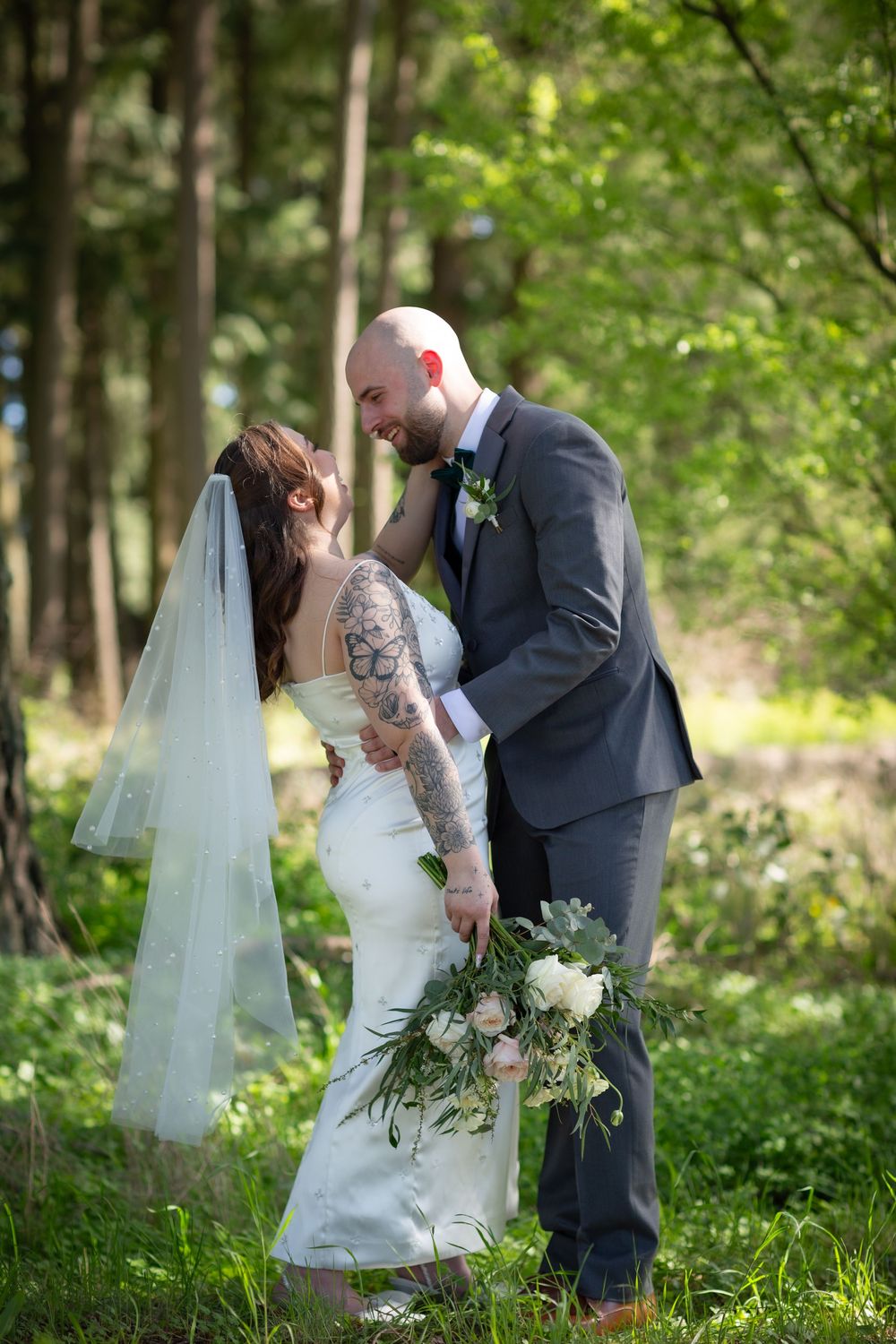 Bride and groom laugh together in lush green forest, groom holding bridal bouquet.