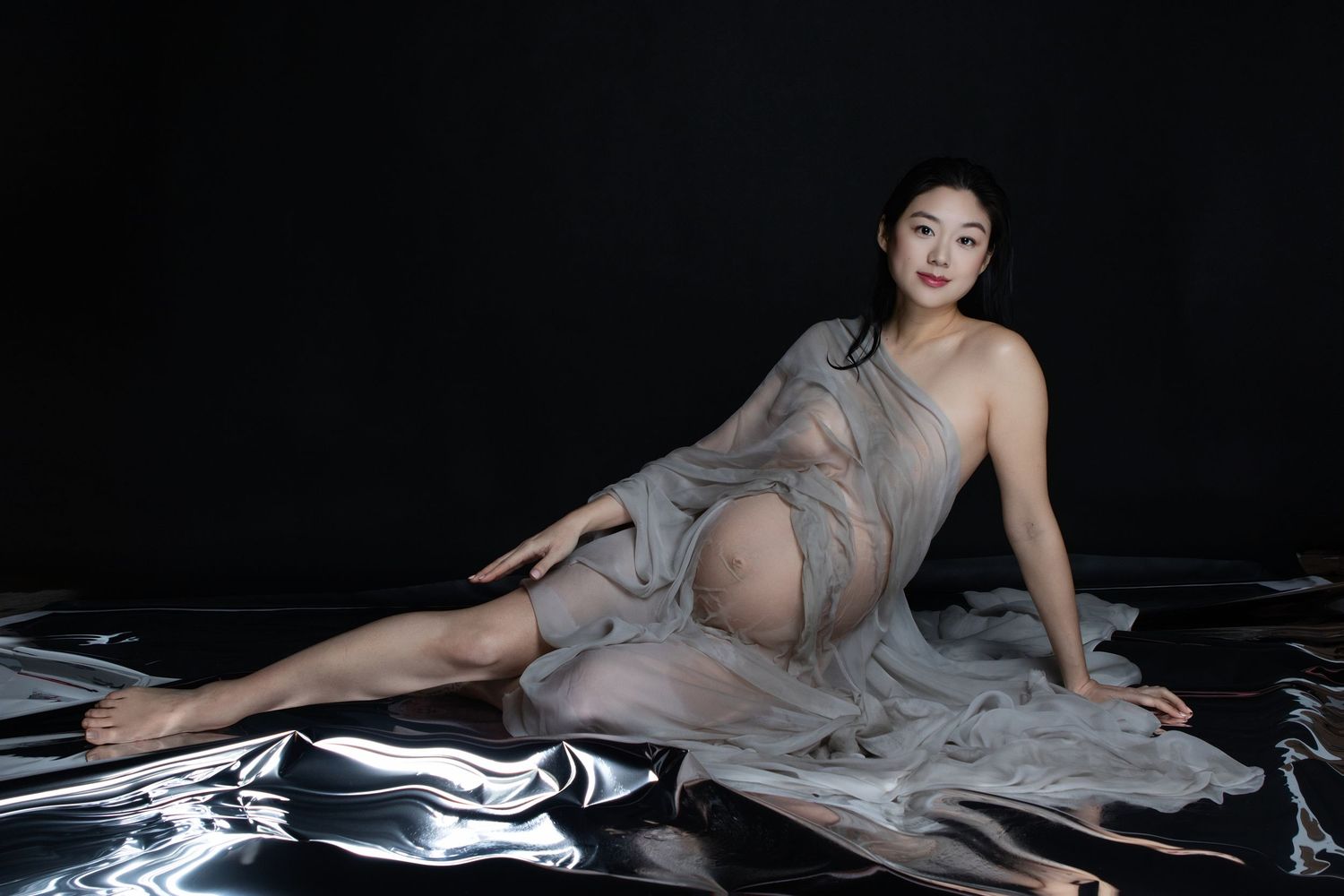 Pregnant woman in sheer gray draped fabric poses gracefully on reflective surface against dark background.