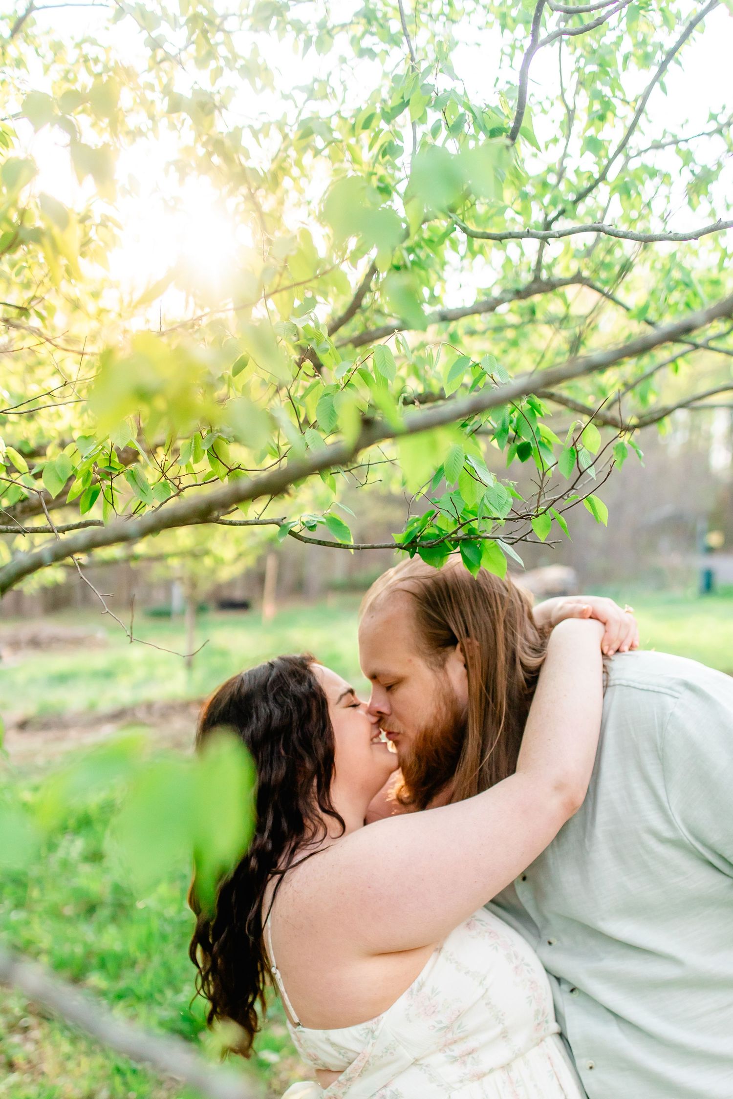 A couple shares a romantic dip kiss under sunlit green tree branches in an outdoor garden setting.
