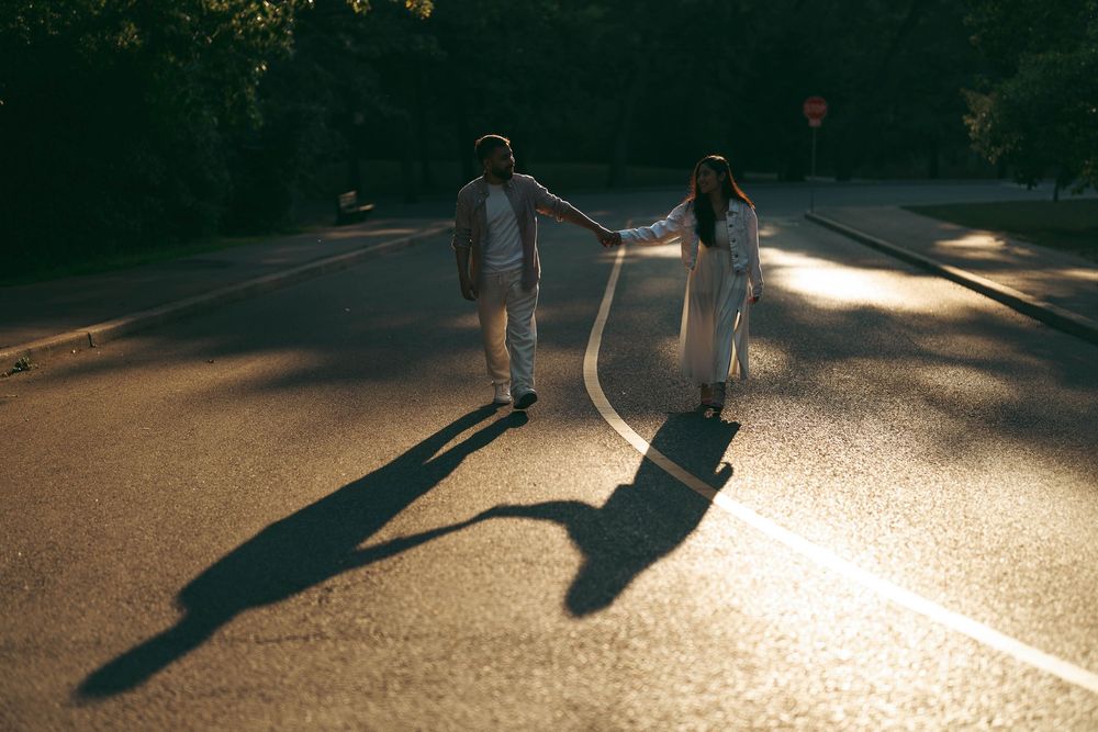 Couple holding hands walking on sunlit road with long shadows, dressed in white, surrounded by lush green trees.