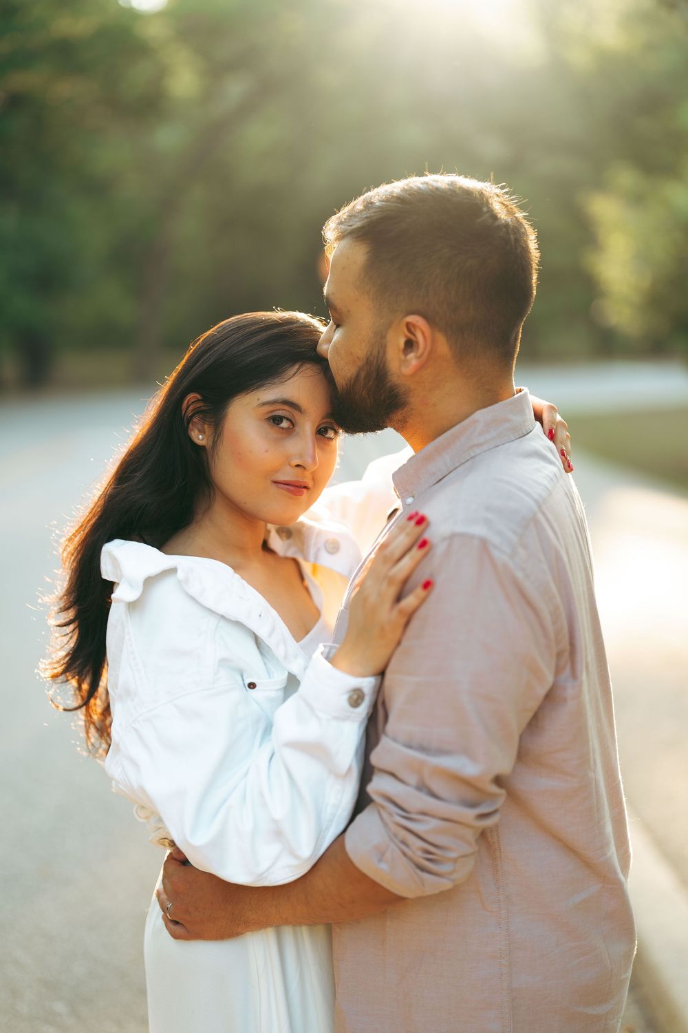 A couple embraces outdoors at sunset, woman in white dress looks at camera while man kisses her head.