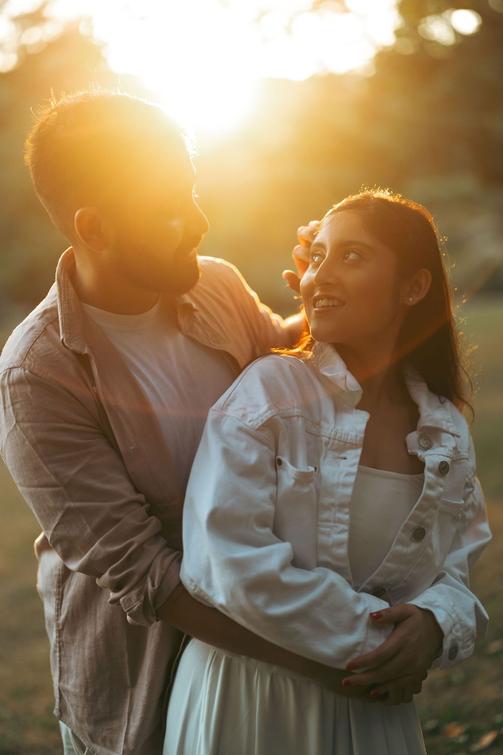 Couple embracing at golden hour sunset, woman in white denim jacket smiling up at man in warm backlit glow.