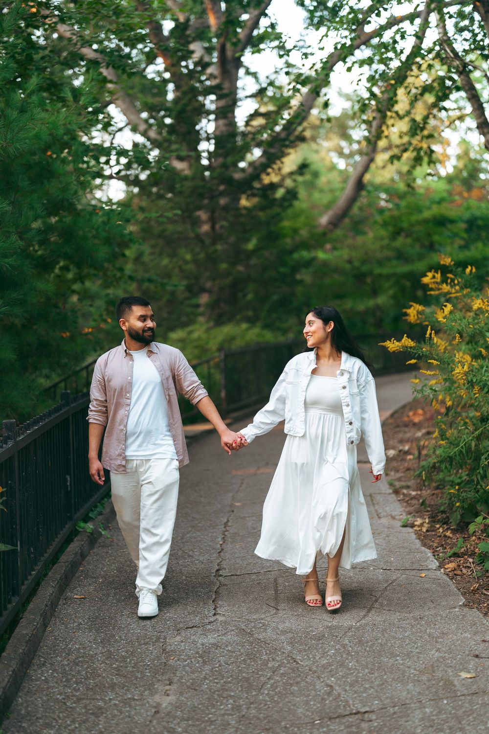 Couple holding hands walking on a park path surrounded by lush green trees, wearing white and pink outfits.