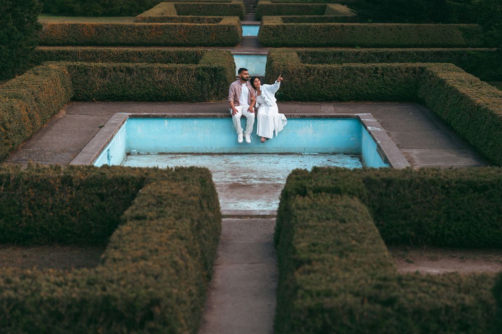 Couple sitting together in a formal hedge maze garden beside a turquoise empty pool.