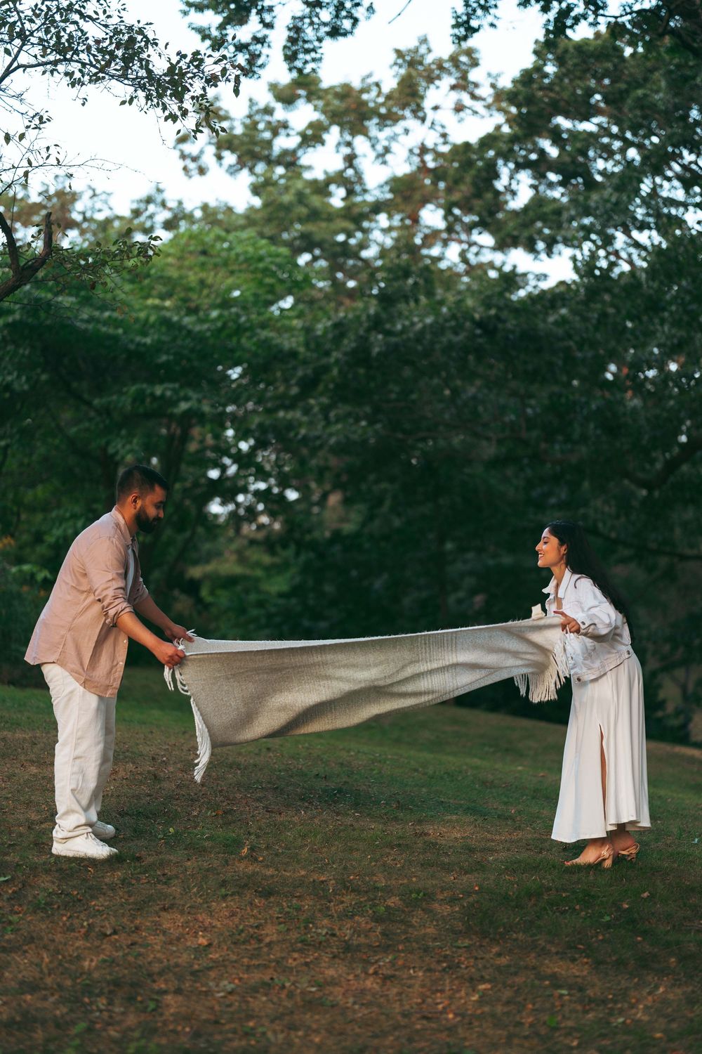 Couple spreading a blanket on grass in a lush green park during a romantic outdoor picnic setup.