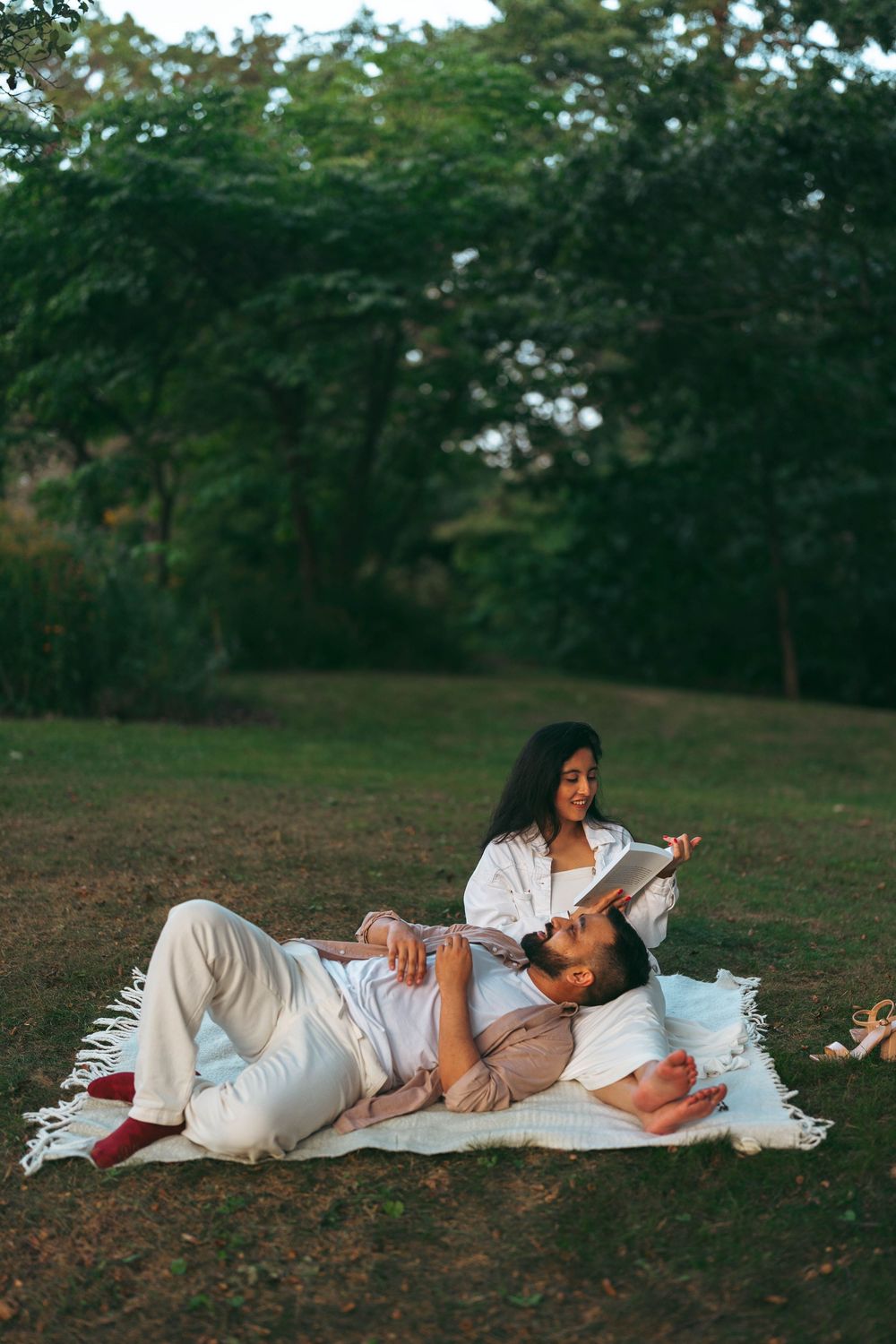 Couple relaxing on a blanket in a lush green park, woman reading a book while man lies with head in her lap.