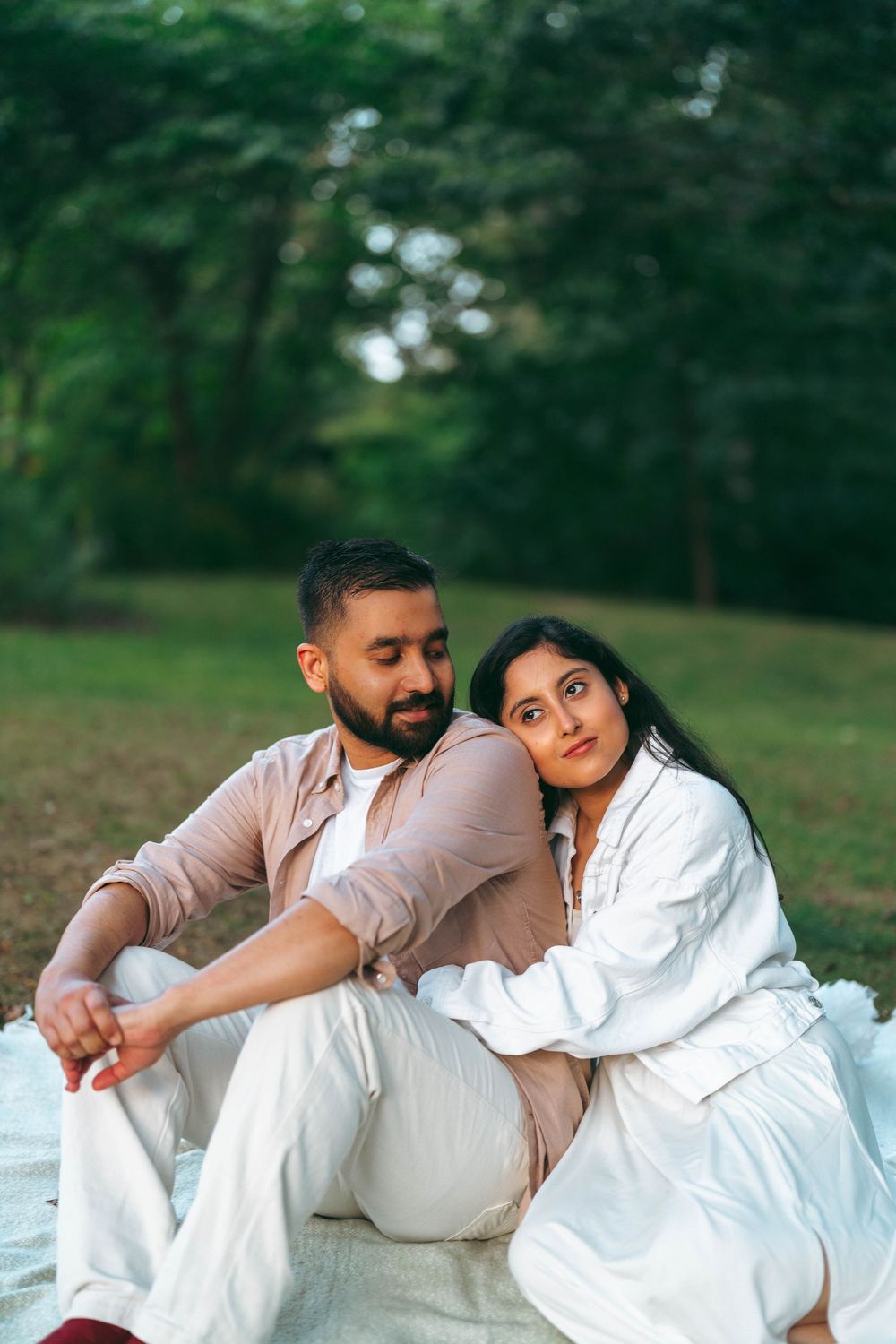A couple sits together on a blanket in a lush green park, woman resting her head on man's shoulder.