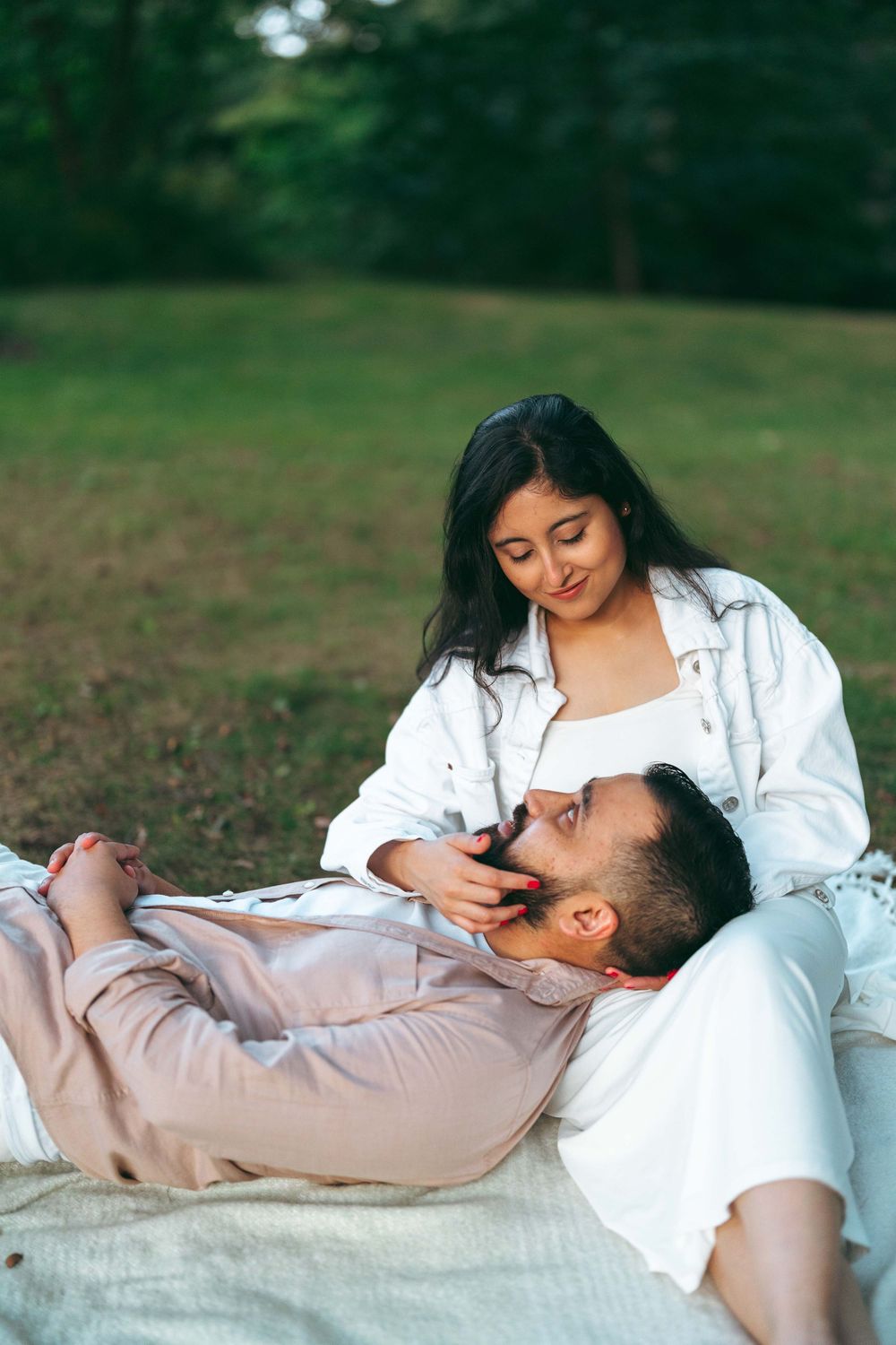 A couple relaxing on a blanket in a park, woman in white jacket gently touching man's beard.