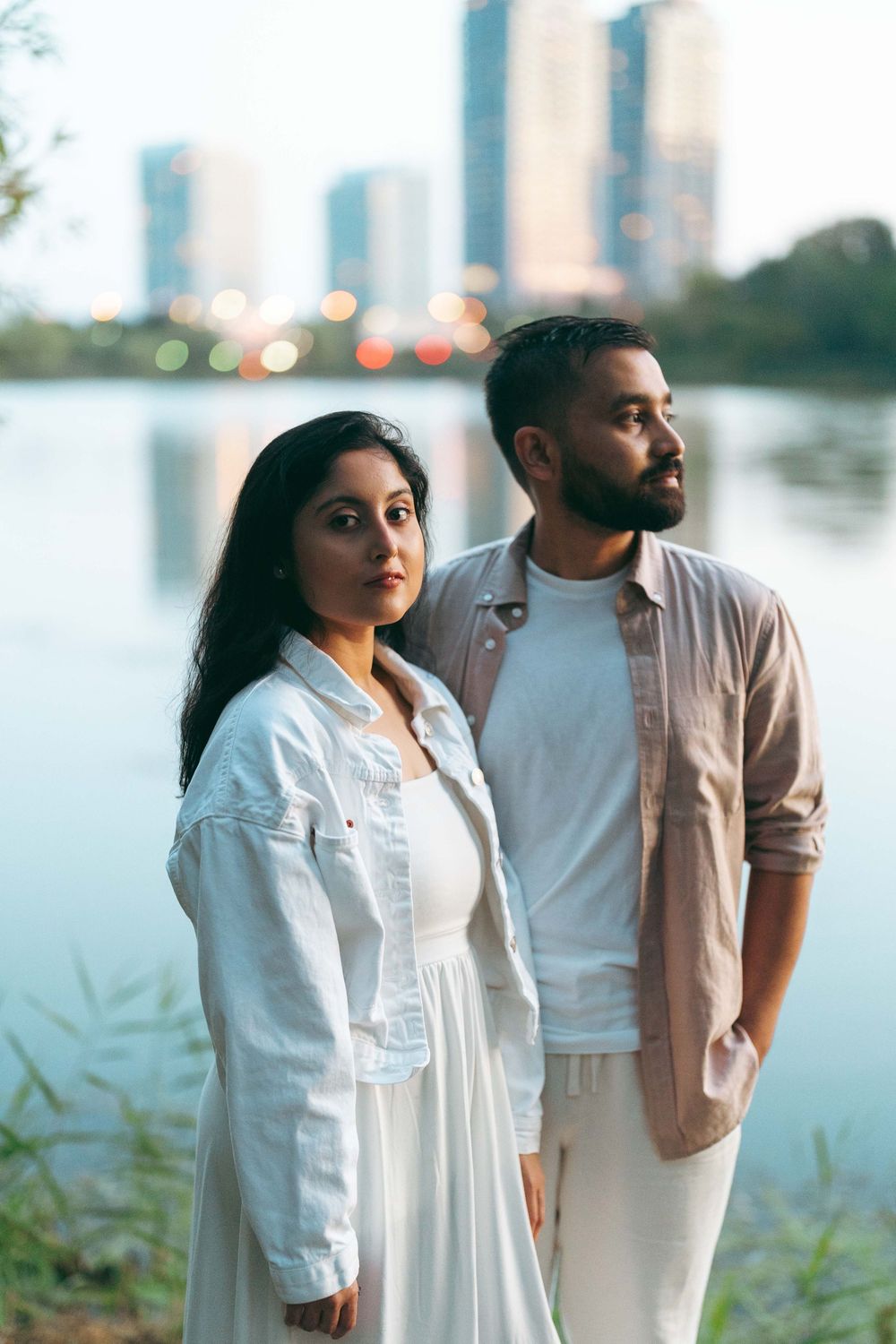 A young South Asian couple poses by a urban lakeside at dusk, woman in white dress and man in beige shirt.