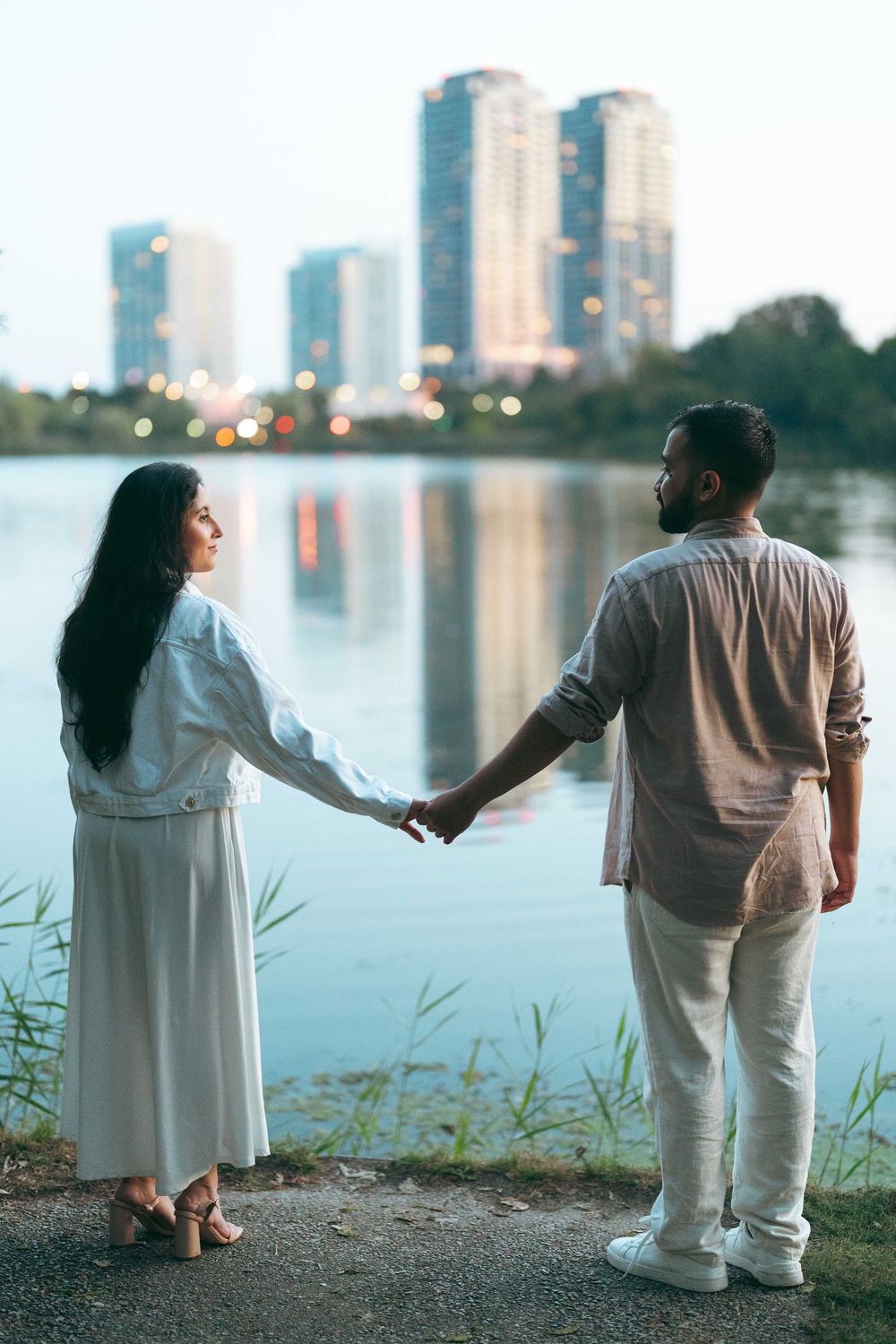 Couple holding hands by a lakeside at dusk with city skyline reflections, woman in white dress, man in casual attire.