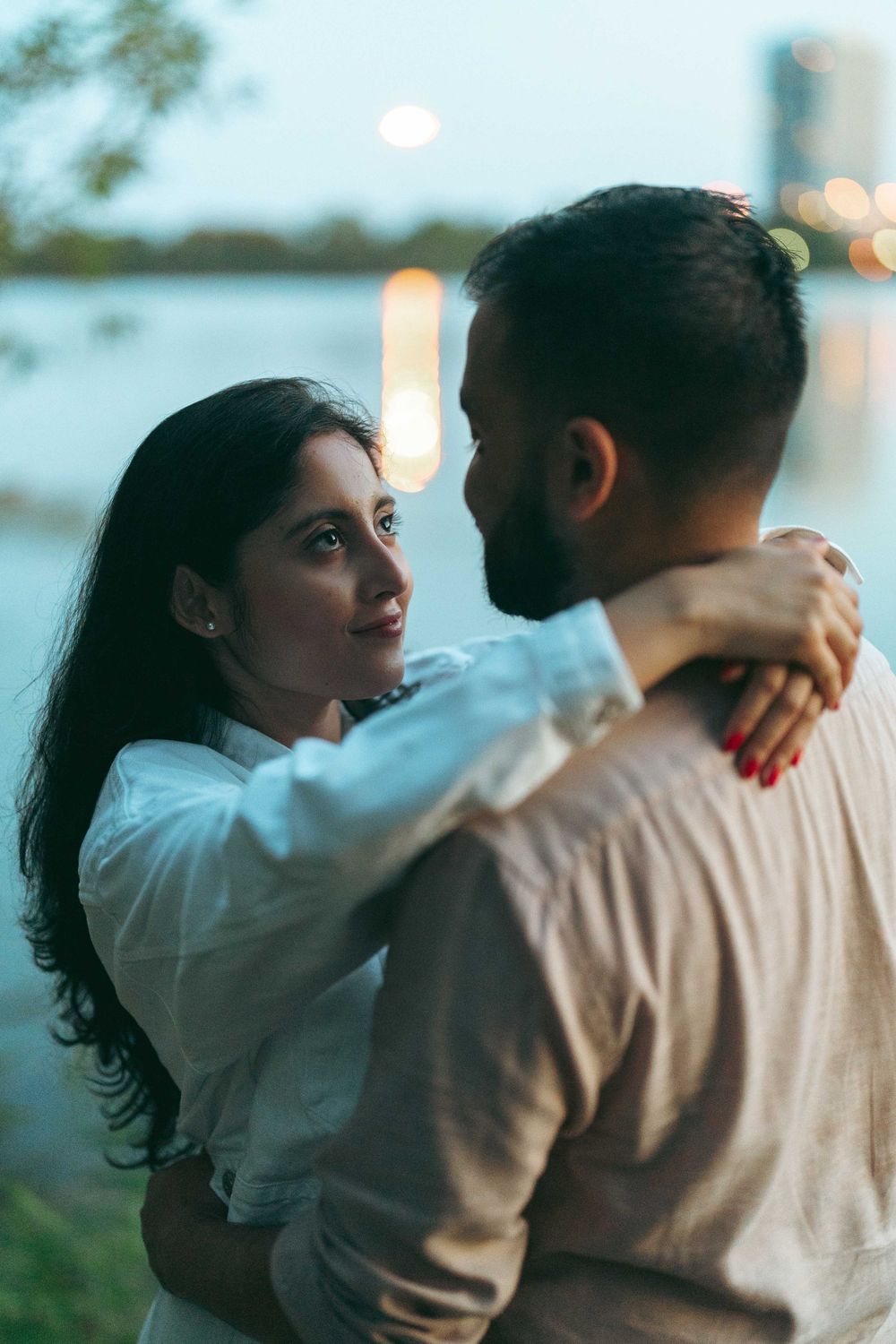 A couple embraces romantically by a lakeside at dusk, woman with red nails gazing lovingly at bearded man.