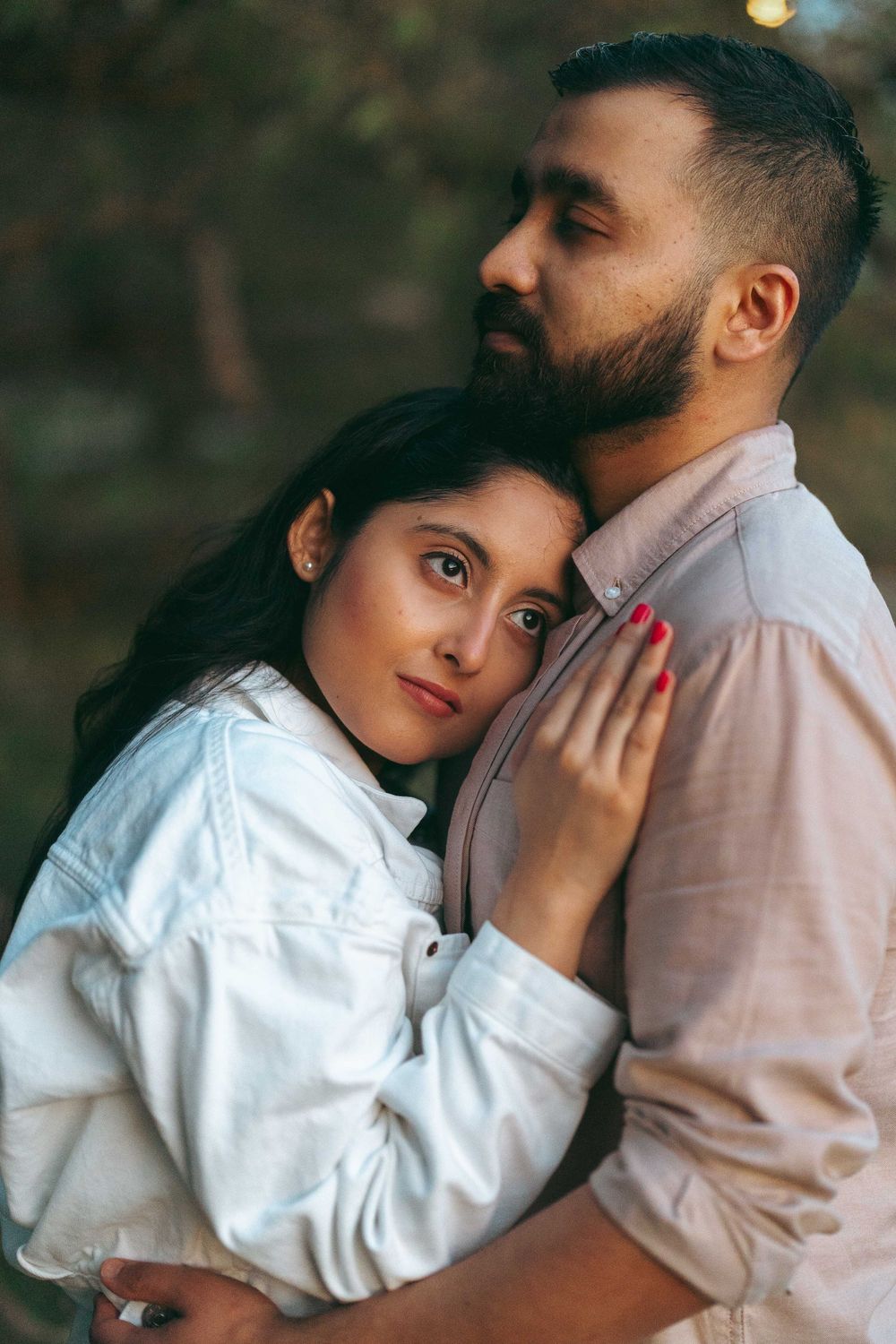 A couple embracing outdoors, woman in white jacket resting head on bearded man's chest in a tender moment.