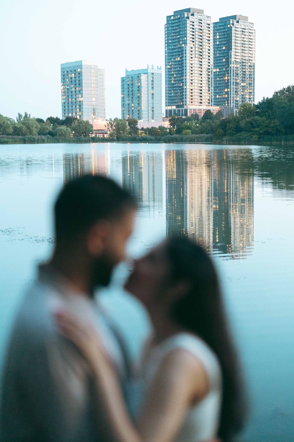 Couple sharing a romantic moment by a reflective urban lake with modern skyscrapers and trees in the background.