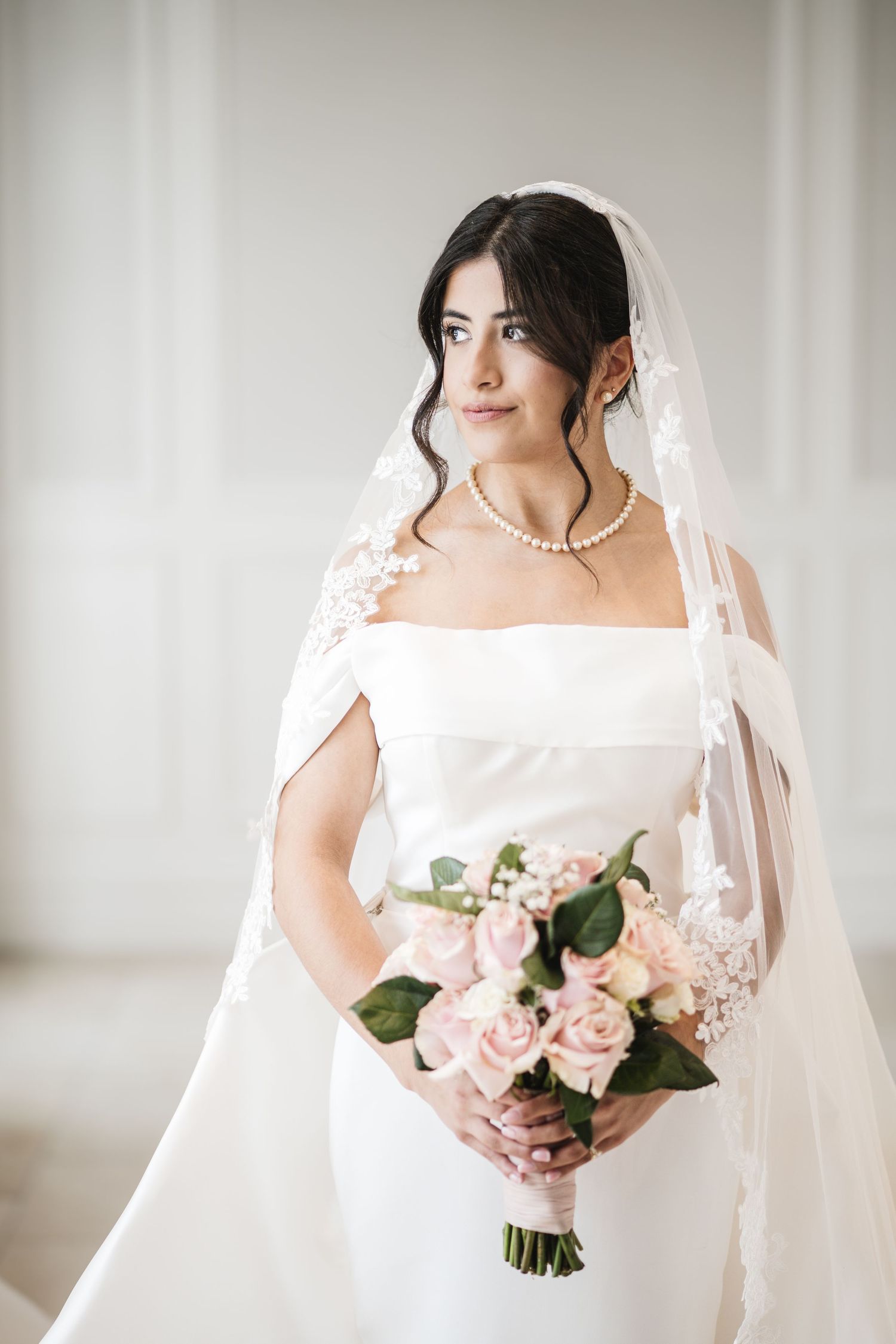Brunette bride in square-neck white gown with lace veil holding a pink rose bouquet with pearl necklace.