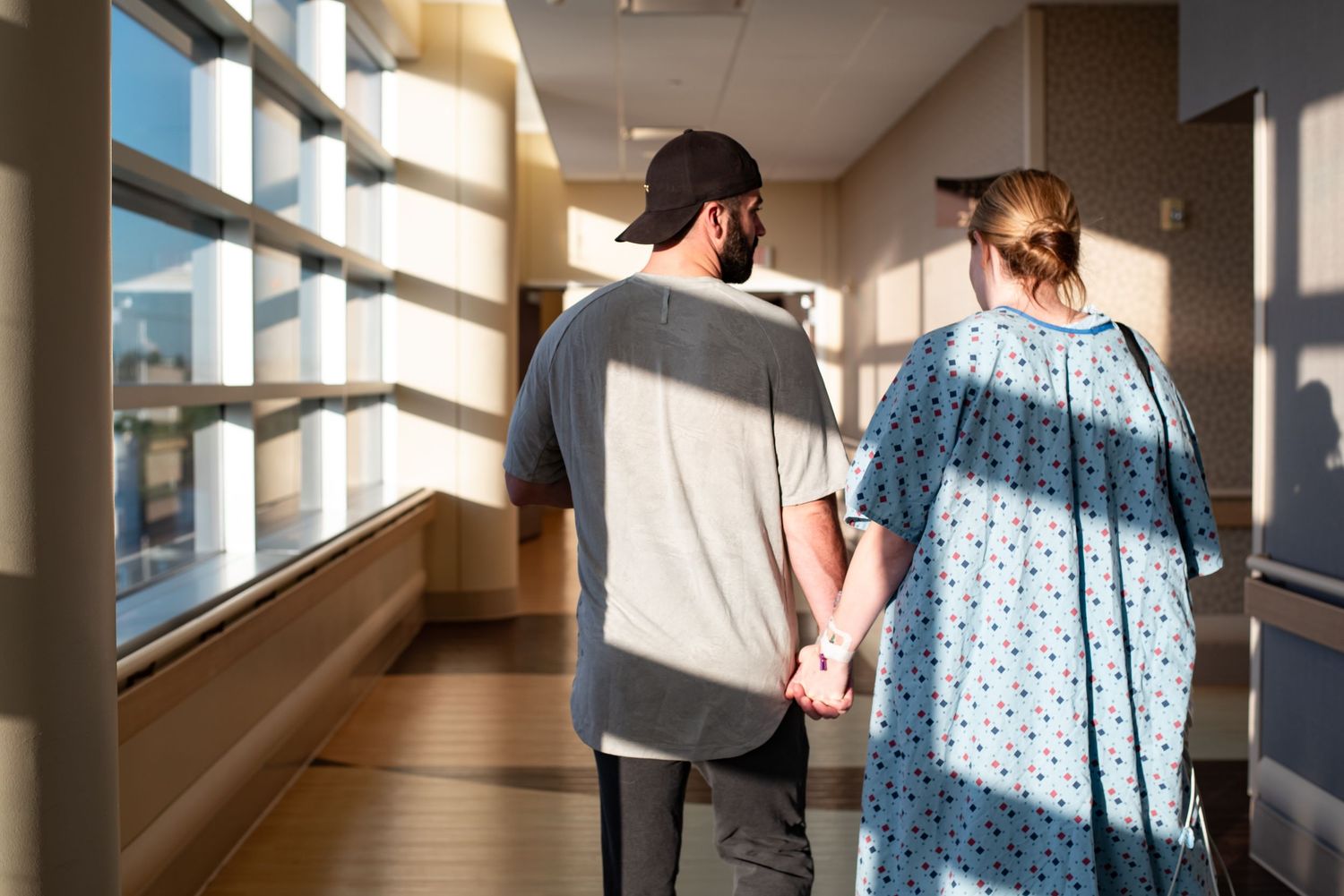 Patient in hospital gown walks with visitor along sunny corridor, holding IV pole during recovery.