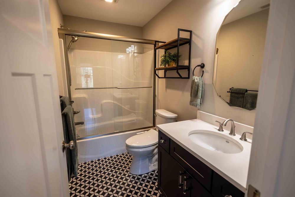Modern bathroom with dark vanity, geometric tile floor, glass shower, and wall-mounted shelving unit.