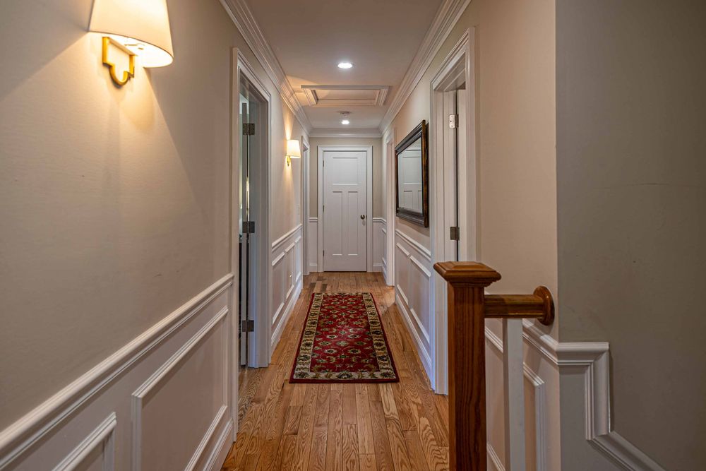 Elegant hallway with wainscoting, hardwood floors, red runner rug, white doors, and wall sconce lighting.
