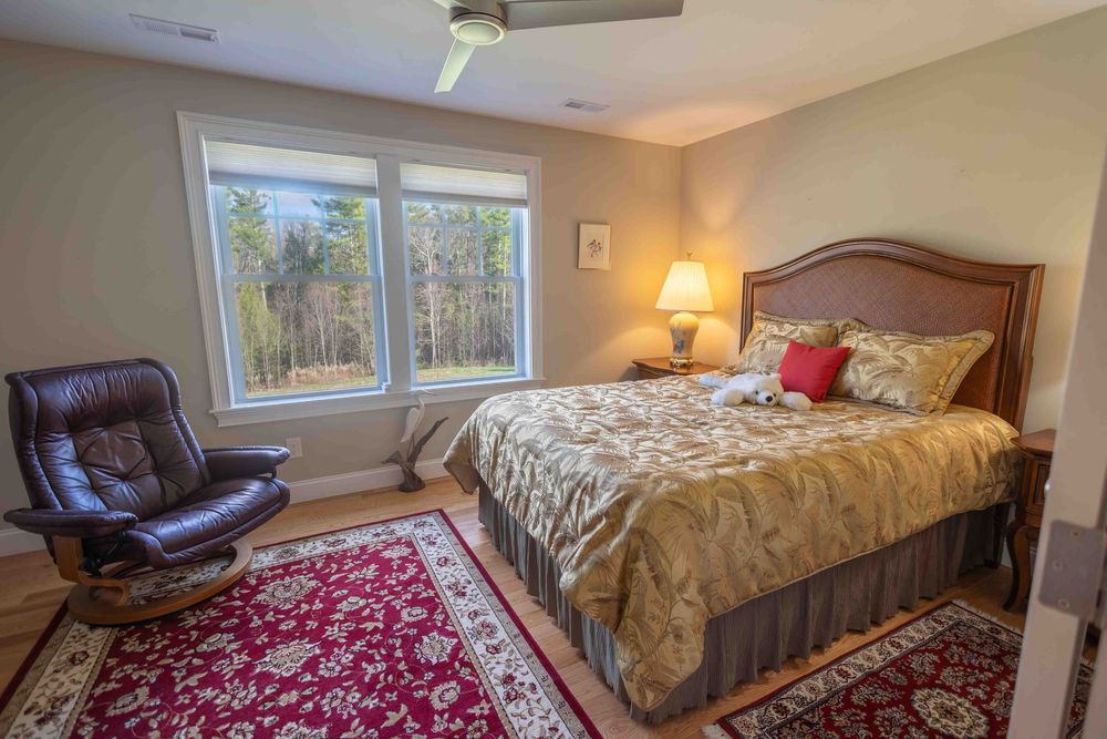 Cozy master bedroom with queen bed, red area rug, leather recliner chair, and large windows overlooking wooded views.