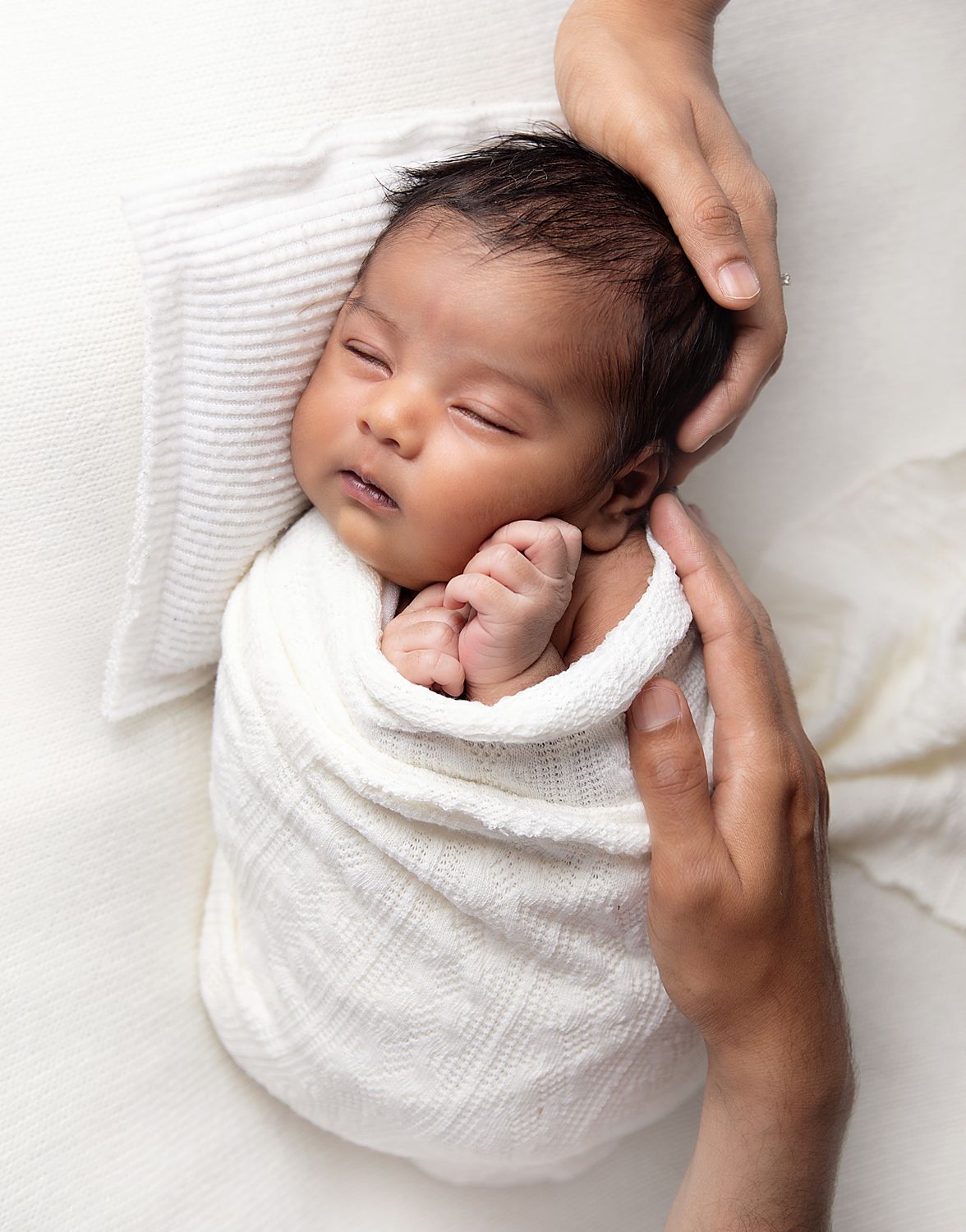 Newborn baby swaddled in white wrap sleeping peacefully while parent's hands gently cradle their head. During a newborn session in Bexley