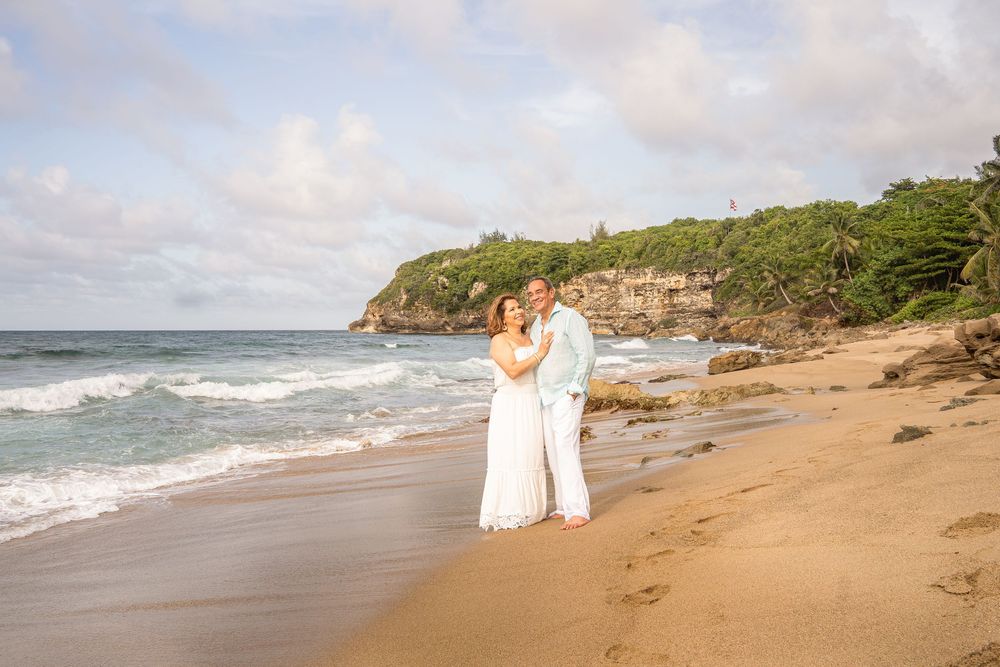 Couple embracing on a sandy tropical beach with ocean waves and lush green cliffs in the background.