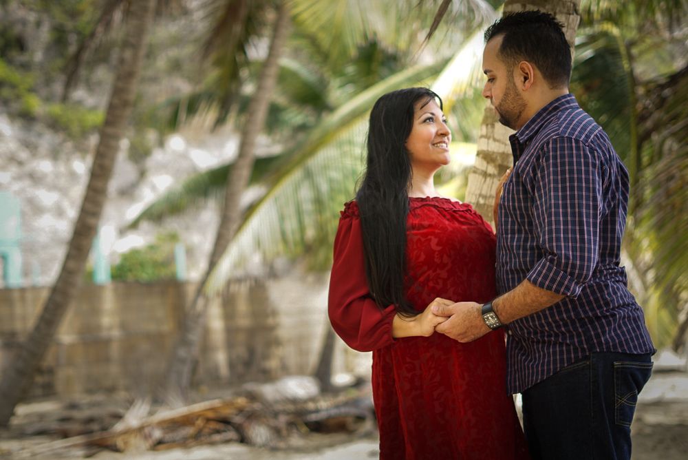 A couple in red and blue attire share a romantic moment on a tropical beach surrounded by palm trees.