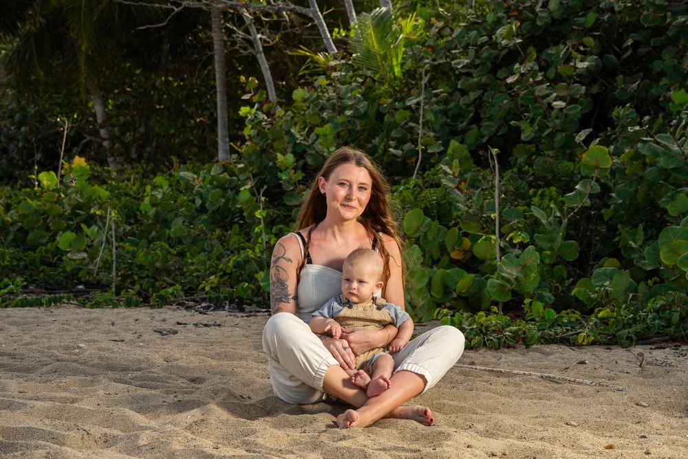 A tattooed mother sits cross-legged on sandy beach holding her baby, with lush tropical greenery behind them.
