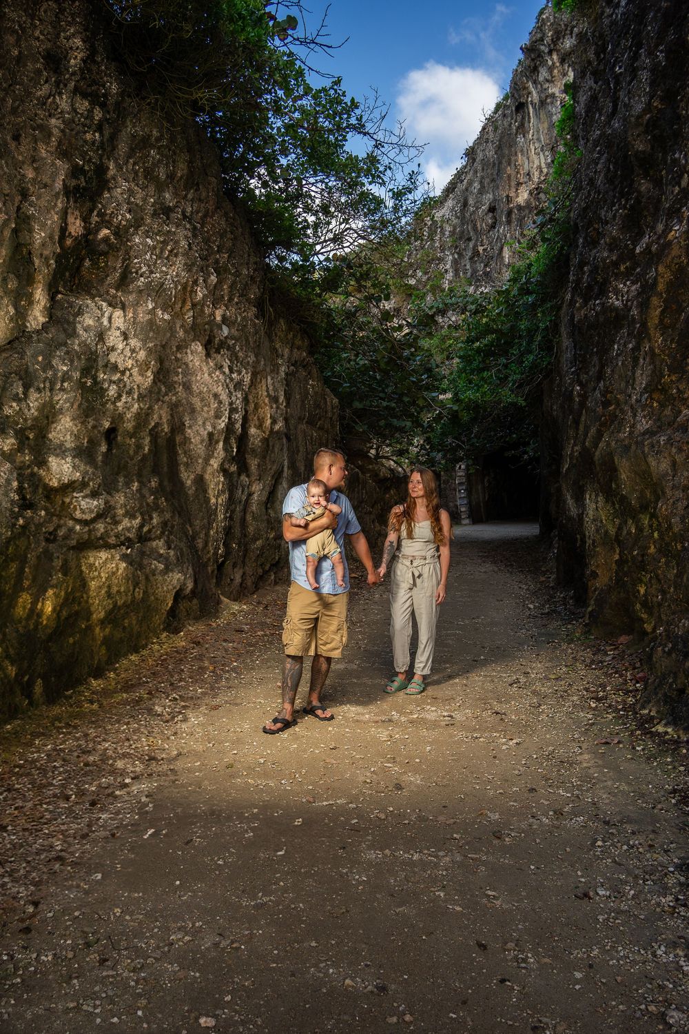 A young family walks hand-in-hand through a narrow rocky canyon path, father holding baby, surrounded by stone walls and greenery.
