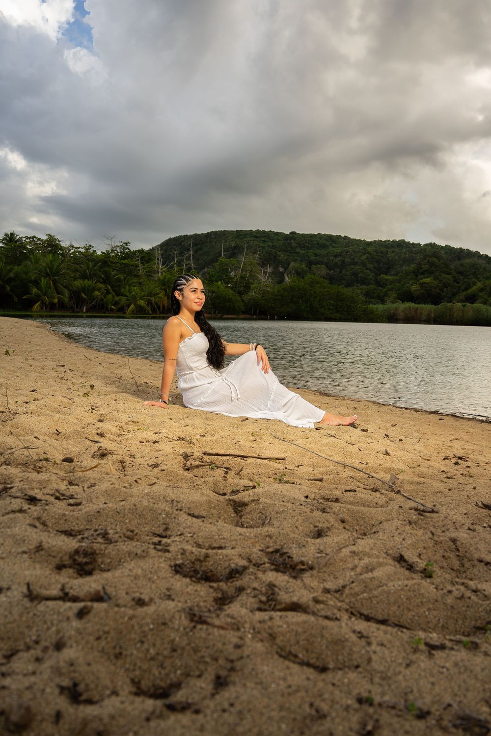 Woman in white dress sitting gracefully on sandy beach with lush green hills and cloudy sky in background.
