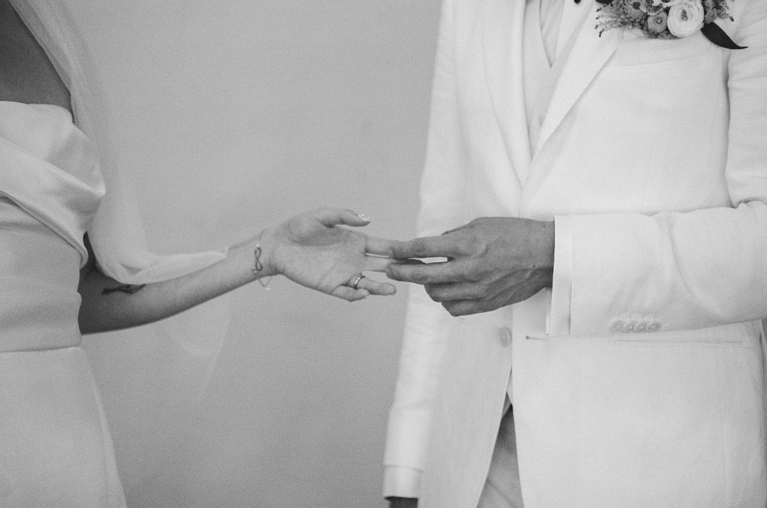 Black and white photo of a couple exchanging wedding rings during a ceremony, groom in white suit.