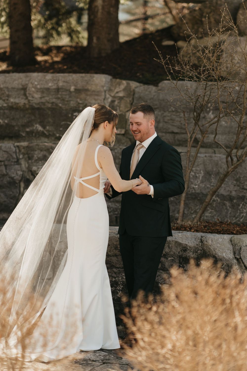 Bride in backless white gown with veil and groom in dark suit share a romantic moment outdoors.