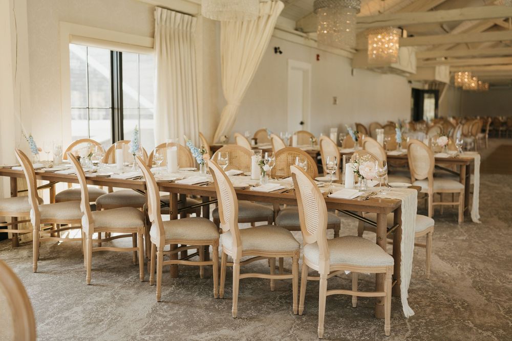 Elegant wedding reception venue with wooden farm tables, French-style chairs, crystal chandeliers, and neutral decor.