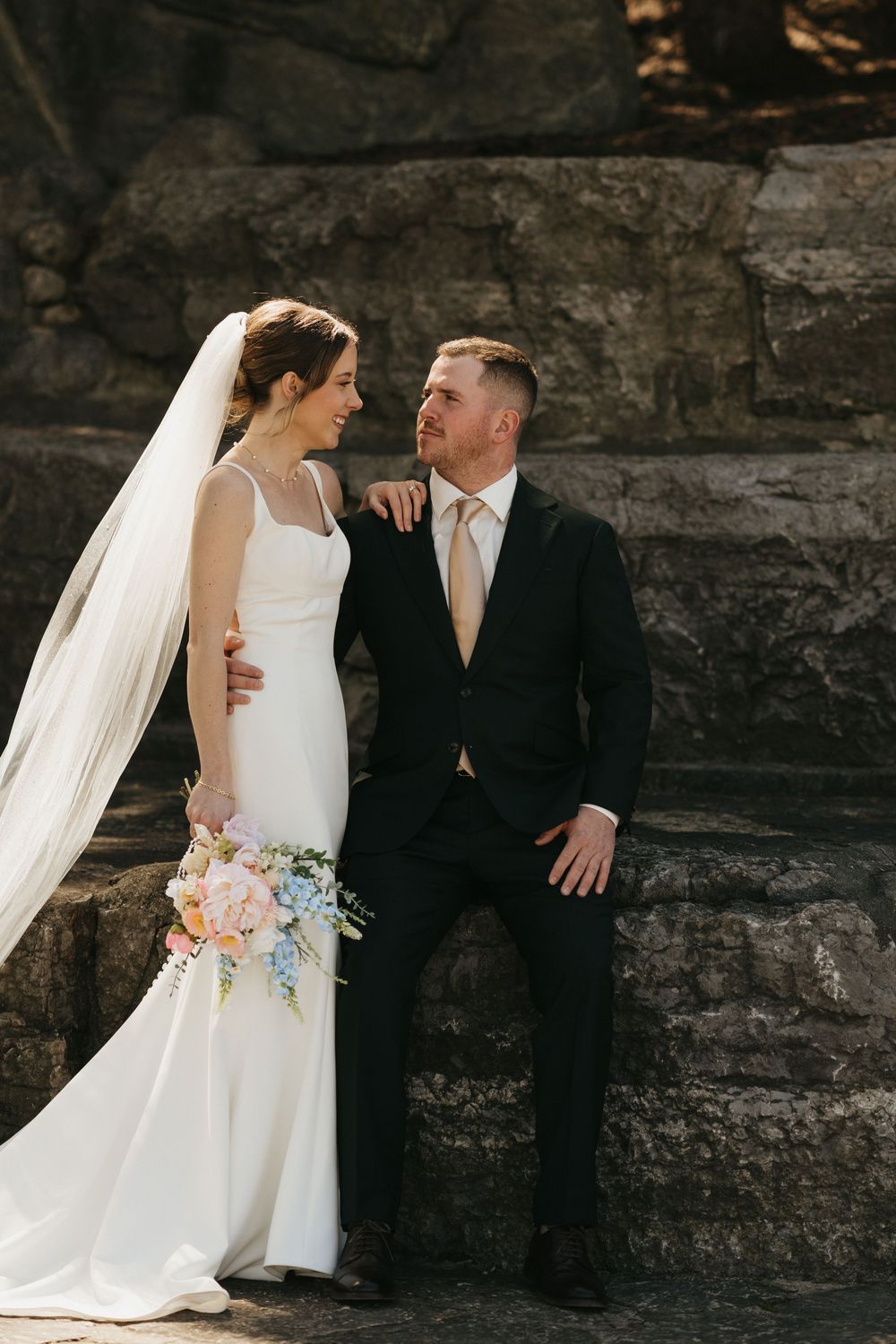 Bride in white gown with veil and groom in black suit pose against stone wall with pastel bouquet.