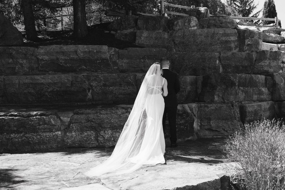Black and white photo of a bride in a long white gown and veil standing before stone steps outdoors.