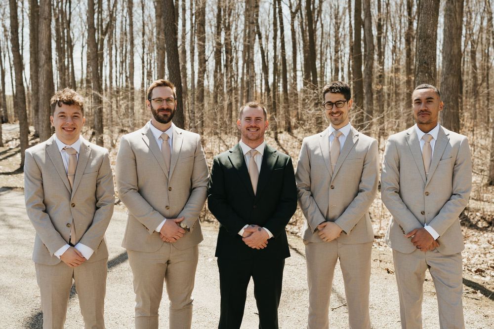 Groom stands with four groomsmen in matching tan suits outdoors in a wooded setting.